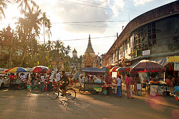 Crowd of people walking in a street market of Yangon, Myanmar. Some guys stand by the stalls, while other walk and ride bicycle. The Shwedagon Pagoda, the main city temple, is visible in the background. Warm sunlight illuminates the scene through palm trees.