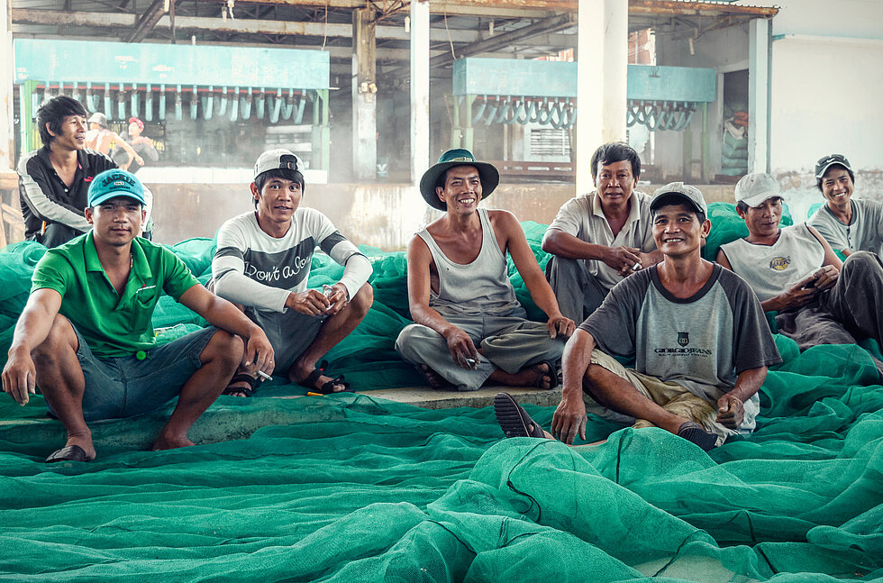 8 male fishers sit at the floor with green fishing net and smile to camera. 