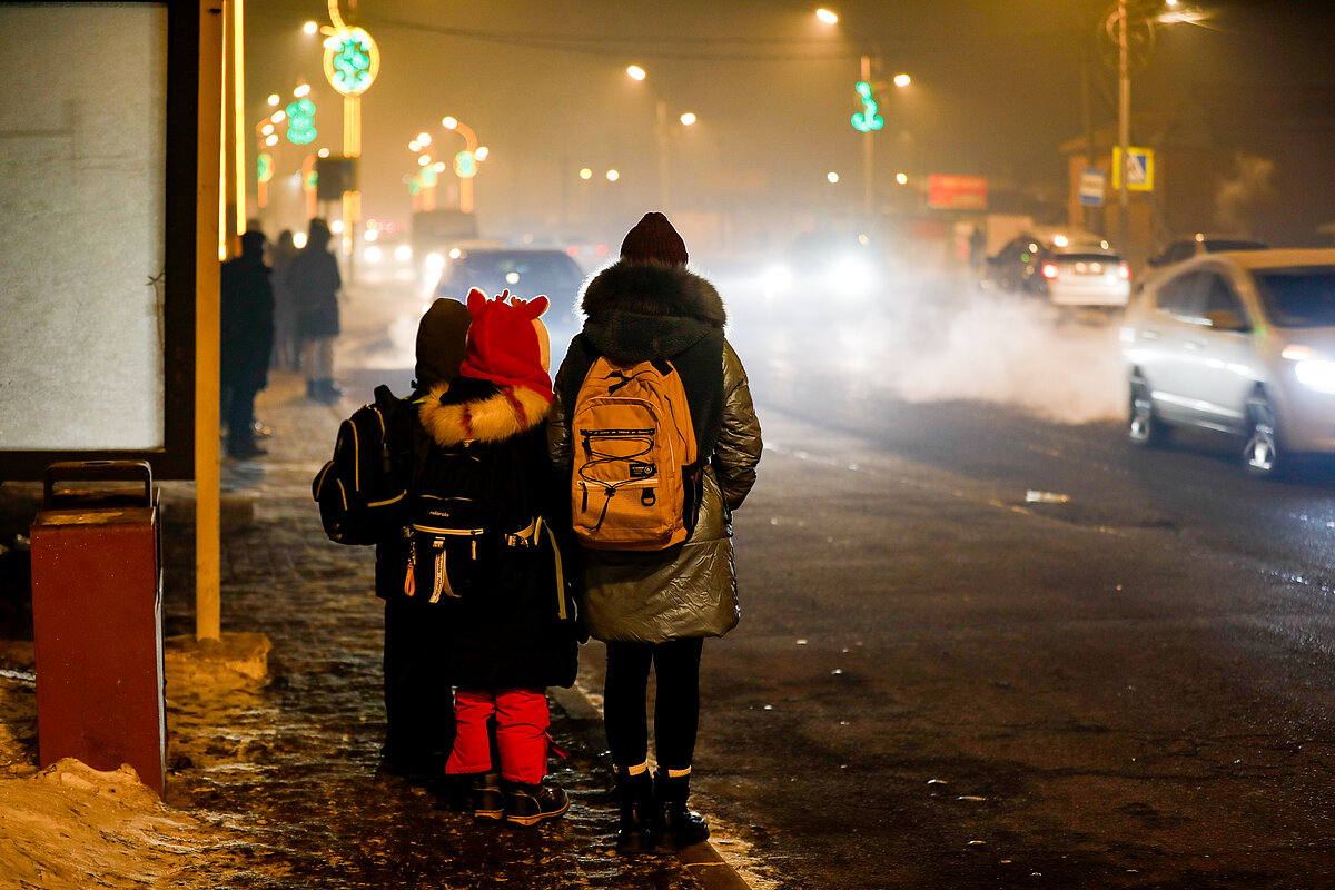 A woman with 2 children standing at streetside at night and looking at the traffic with air pollution