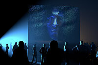 an image of a dark room filled with crowd and a huge human face at the background
