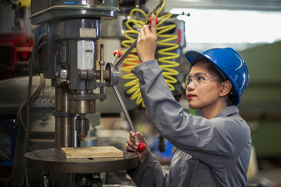 An Asian woman wearing blue helmet and protective glasses uses an industrial drill in a plant.